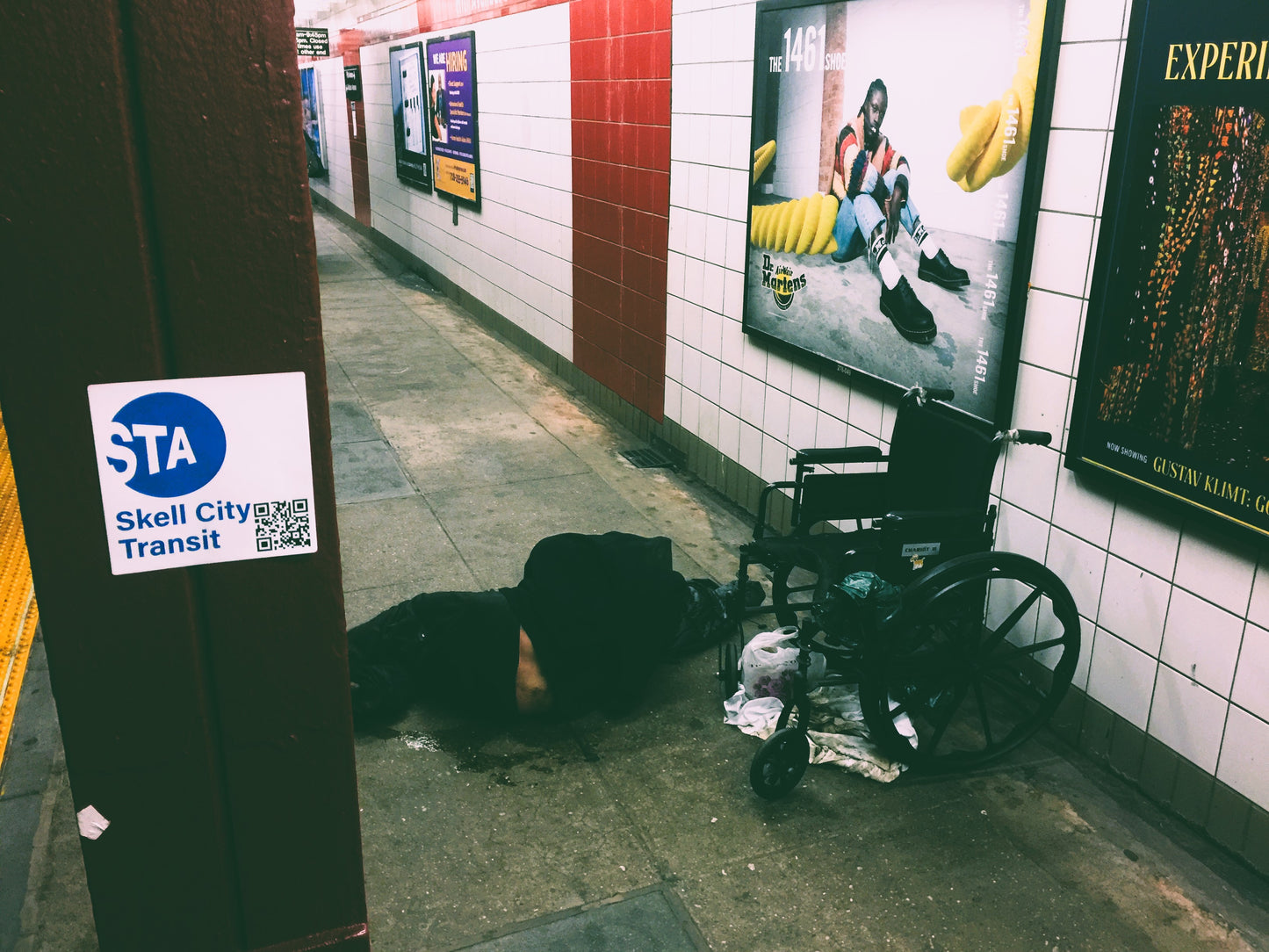 MTA station sticker from skell city, on a red subway platform beam with a soiled homeless man sleeping on the ground covered in urine, next to his wheelchair.