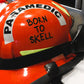Paramedic Orange Bunker Gear Helmet On a Stretcher, which has a Clear Born To Skell Sticker On the Side of the helmet.