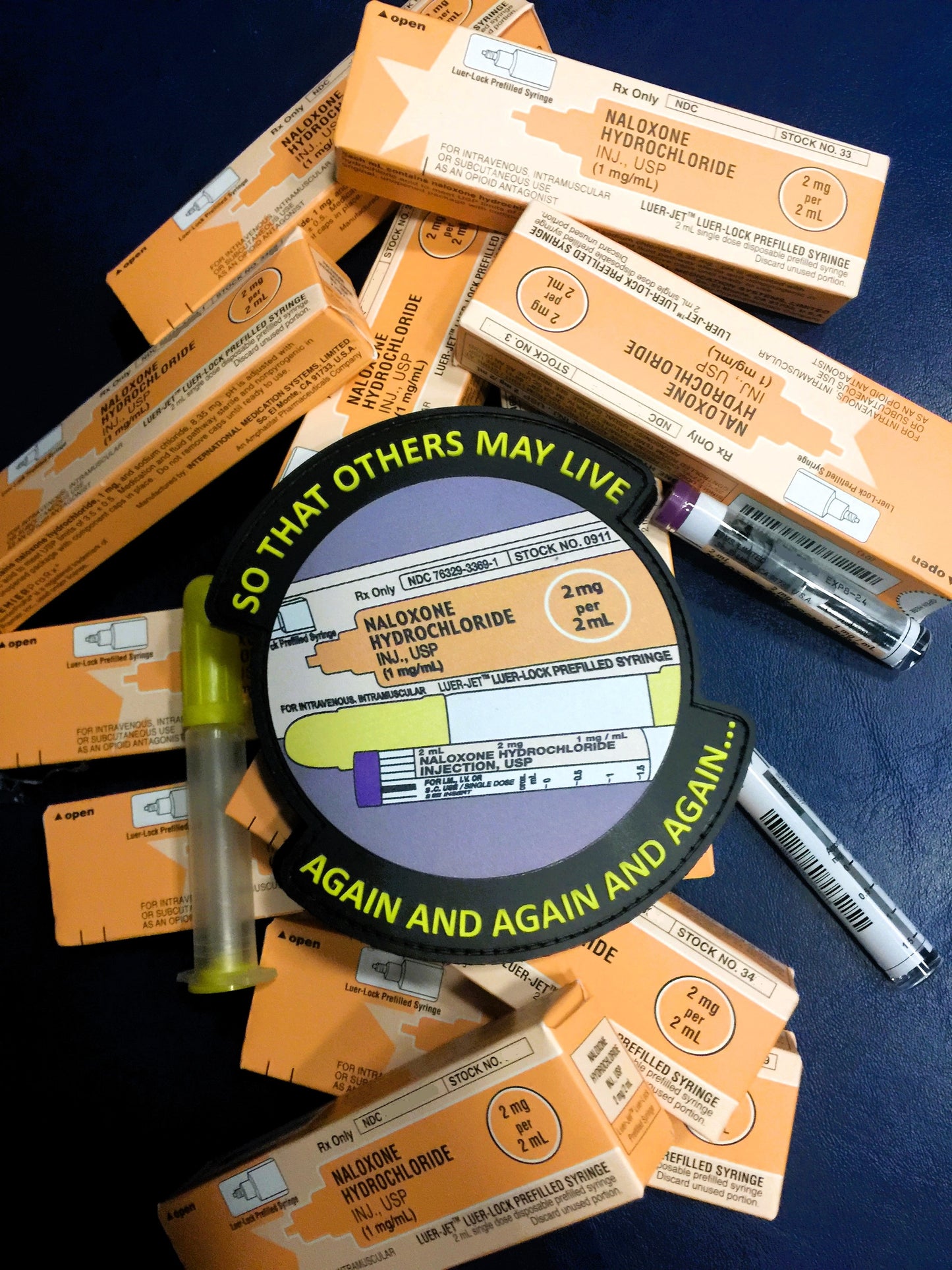 So that others may live Narcan PVC Velcro patch from skell city, lying on top of naloxone hydrochloride boxes and purple vials.