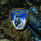 EMS patch from skell city lying on dirty wet leaf covered ground, next to garbage.
